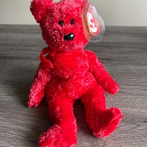 May include: A bright red plush teddy bear with a black nose and eyes, and a red ribbon tied around its neck. The bear has a clear tag attached to its ear. The toy is sitting on a wooden surface.