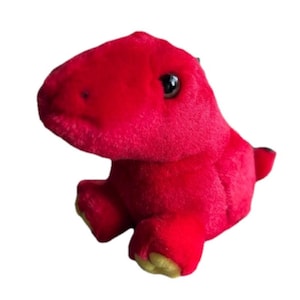 Może przedstawiać: Pluszowa czerwona zabawka dinozaura z czarnymi oczami i żółto-zielonymi stopami. Pluszak siedzi, z lekko pochyloną głową. Zabawka wykonana jest z miękkiego, puszystego materiału.
