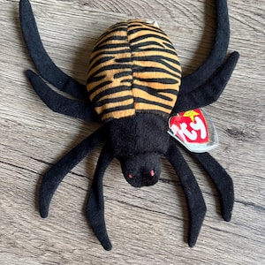 Può includere: Un peluche a forma di ragno con un corpo a strisce arancioni e nere e lunghe zampe nere. Il ragno ha gli occhi rossi e un'etichetta attaccata. Il giocattolo è progettato per assomigliare a un ragno.