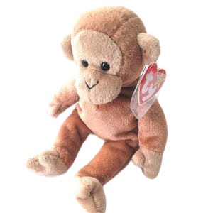 May include: A plush monkey toy with brown and tan fur. The monkey has a light brown face, ears, hands, and feet. It has black eyes and a stitched smile. A red and white tag is attached to the monkey's ear.