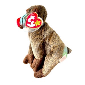 May include: A brown plush baboon toy with a red, white, and blue nose and a red heart-shaped tag. The baboon has brown and tan fur, brown feet, and a small green patch on its rear. The tag reads "Beanie Baby".