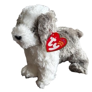 Puede incluir: Un juguete de peluche con forma de perro con pelaje blanco y gris. El juguete tiene una nariz negra y una etiqueta roja en forma de corazón que dice "Beanie Babies". El perro está de pie sobre sus cuatro patas.