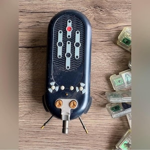 Puede incluir: Un dispositivo electrónico vintage azul oscuro con múltiples botones y deslizadores. El dispositivo tiene una serie de pequeñas luces y una luz roja central. Dos estructuras doradas en forma de antena se extienden desde la parte inferior. Se ven varios cartuchos de juego de plástico transparente.