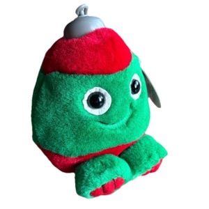 May include: A plush Christmas ornament in the shape of a smiling green creature with large black eyes and a red cap and feet. The ornament has a silver top and a tag attached.