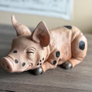 以下が含まれることがあります： リラックスしたポーズのセラミック製の豚の置物。目を閉じ、穏やかな笑顔を浮かべている。豚は薄いピンク色で、黒い斑点と灰色の蹄がある。置物は木製の表面に置かれている。