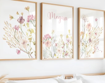 May include: Three framed floral art prints with watercolor flowers in shades of pink, purple, and yellow. The middle print has the name "Megan" in pink script. The frames are light brown, and the prints are displayed above a bed.