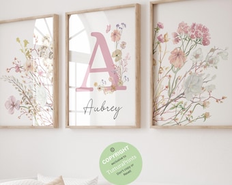May include: Three framed floral art prints. The center print features a pink letter "A" with the name "Aubrey" below. The other two prints display watercolor floral arrangements in soft pinks, greens, and whites. The frames are light wood.