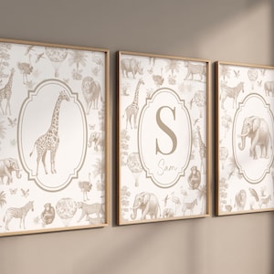 May include: Three framed wall art prints featuring a beige and white safari animal pattern. The left print shows a giraffe, the center print displays the letter S and the name Sam, and the right print depicts an elephant, all within oval frames.