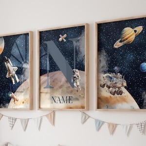 May include: Three framed prints with a space theme. The prints feature a rocket, an astronaut, a planet, and a letter N. The middle print has the text "NAME" below the letter N.