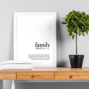 May include: A framed print with the word "family" in a minimalist style, accompanied by a definition. A small potted plant and a folded white towel with black trim sit on a wooden desk with drawers. The background is a light gray wall.