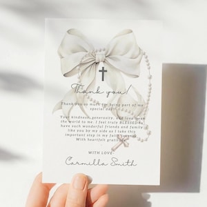 May include: A white card featuring a bow, cross, and rosary bead design. The card includes the text "Thank you!" and a personalized message, suitable for a religious occasion. The card is held by a person.