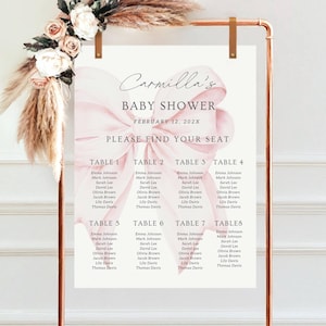 May include: A copper-coloured metal frame displays a baby shower seating chart. The chart features a pink bow design and the text "Carmilla's BABY SHOWER" with the date. Floral and pampas grass accents adorn the top of the frame.