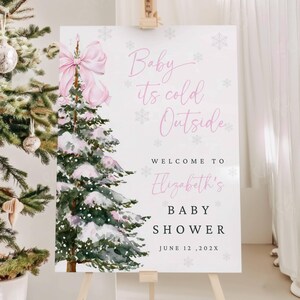 May include: A white sign on a wooden easel with the text "Baby it's cold Outside" and "Welcome to Elizabeth's Baby Shower June 12, 202X". The sign features a pink bow and a watercolor Christmas tree design. A decorated Christmas tree is in the background.