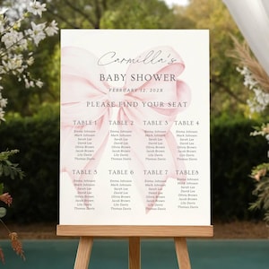 May include: A baby shower seating plan displayed on a wooden easel. The sign features a pink watercolour ribbon design and the text "Carmilla's BABY SHOWER FEBRUARY 12, 202X PLEASE FIND YOUR SEAT". Table assignments are listed.