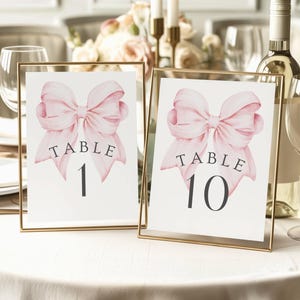 May include: Two framed table number cards with gold frames. Each card features a watercolor pink bow and the word "TABLE" above a number. Table 1 and Table 10 are displayed on a white tablecloth.