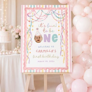 Pode incluir: Uma placa retangular de boas-vindas de aniversário num cavalete de madeira com borda de listras pastel, guirlandas em aquarela e estrelas. O texto diz: it's fun to be one, WELCOME TO CARMILLA'S first birthday, MARCH 10, 202X, com uma foto.