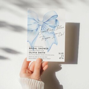 May include: A blue watercolor bow with the text "Something Blue Before I Do!" on a white background. The text below the bow reads "Please join us for a Bridal Shower honoring bride-to-be Olivia Smith 123 Little Peanut Road Chicago 60556 RSVP to Jane at 123-456-7890 Registered at Amazon & Target MAR 24 4PM".