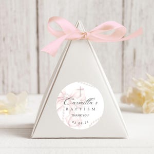 Könnte beinhalten: Weiße dreieckige Geschenkbox mit einem rosa Satinband. Ein runder Aufkleber mit einer rosa Schleife, einem Kreuz und dem Text "Carmillas Taufe Danke 03.08.21" befindet sich auf der Vorderseite der Box.