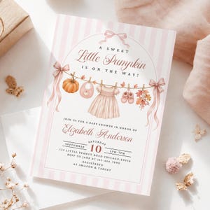 May include: A baby shower invitation card with light pink vertical stripes, featuring a clothesline illustration with a pumpkin, bib, dress, and booties. Text reads: A SWEET Little Pumpkin IS ON THE WAY! JOIN US FOR A BABY SHOWER IN HONOR OF Elizabeth Anderson SATURDAY SEPTEMBER 10 2PM-7PM 123 LITTLE PEANUT ROAD CHICAGO, 60556 RSVP TO JANE AT 123-456-7890 REGISTERED AT AMAZON & TARGET.