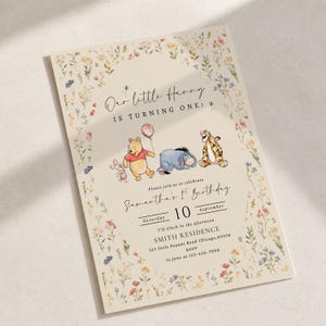 Puede incluir: Una tarjeta de invitación de cumpleaños color crema con un borde floral en acuarela e ilustraciones de Winnie the Pooh, Piglet, Eeyore y Tigger. El texto dice Our little Hunny IS TURNING ONE, Samantha's 1st Birthday, 10 de septiembre y detalles.