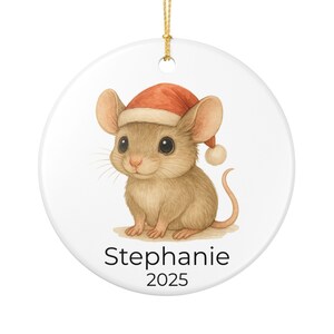 Può includere: Ornamento natalizio rotondo e bianco con un'illustrazione ad acquerello di un simpatico topo con un cappello da Babbo Natale. Il topo è marrone chiaro con grandi occhi e baffi. Il nome "Stephanie" e l'anno "2025" sono stampati sotto. Un cordino dorato è attaccato per appenderlo.