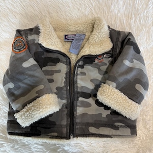 Puede incluir: Chaqueta con estampado de camuflaje con cuello y puños de sherpa color crema. La chaqueta tiene cierre de cremallera y un parche en la manga izquierda que dice "Junior Pilot Association". Chaqueta para niño.