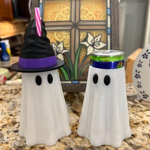 May include: Two white ghost-shaped beverage can coolers. The left ghost wears a black witch hat with a purple band and a pink and white striped straw. The right ghost holds a green and blue aluminum soda can. Both have two black oval eyes.