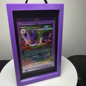 Peut inclure: Une carte Pok&eacute;mon encadr&eacute;e avec Mega Gengar EX. La carte est pr&eacute;sent&eacute;e dans un cadre violet avec un int&eacute;rieur noir et une poign&eacute;e noire. L'illustration de la carte montre une cr&eacute;ature violette et noire avec le texte "Mega Gengar EX" et "350".