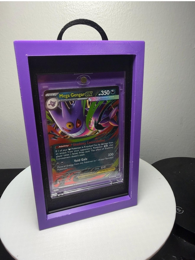 Peut inclure: Une carte Pok&eacute;mon encadr&eacute;e avec Mega Gengar EX. La carte est pr&eacute;sent&eacute;e dans un cadre violet avec un int&eacute;rieur noir et une poign&eacute;e noire. L'illustration de la carte montre une cr&eacute;ature violette et noire avec le texte "Mega Gengar EX" et "350".