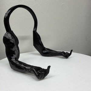 May include: A black, abstract sculpture resembling a stylized human form. The piece features a curved top section and two arm-like extensions with hand details. The sculpture is set against a plain white background.