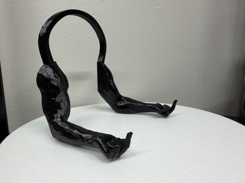 May include: A black, abstract sculpture resembling a stylized human form. The piece features a curved top section and two arm-like extensions with hand details. The sculpture is set against a plain white background.