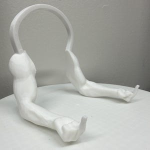 May include: A white, 3D-printed headphone stand shaped like a pair of muscular arms. The arms are extended, with the hands forming a gesture. The stand has a curved top to hold headphones. The product is designed to be placed on a flat surface.