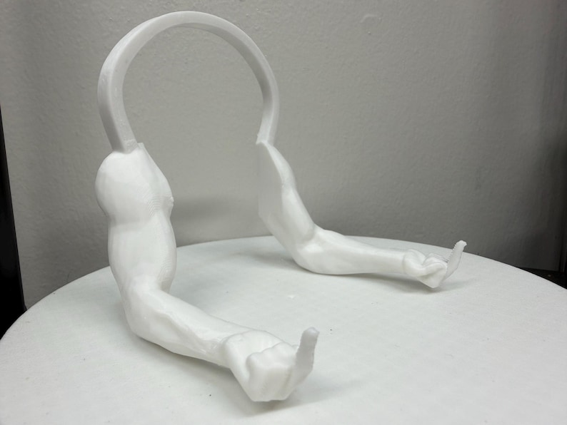 May include: A white, 3D-printed headphone stand shaped like a pair of muscular arms. The arms are extended, with the hands forming a gesture. The stand has a curved top to hold headphones. The product is designed to be placed on a flat surface.
