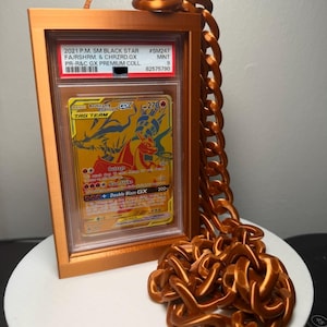 Peut inclure: Une carte &agrave; collectionner encadr&eacute;e avec un design dor&eacute; et orange, avec le texte "Reshiram & Charizard GX TAG TEAM". Le cadre et la cha&icirc;ne sont de couleur cuivre m&eacute;tallique. La carte est not&eacute;e et &eacute;tiquet&eacute;e avec le texte "2021 P.M. SM BLACK STAR".