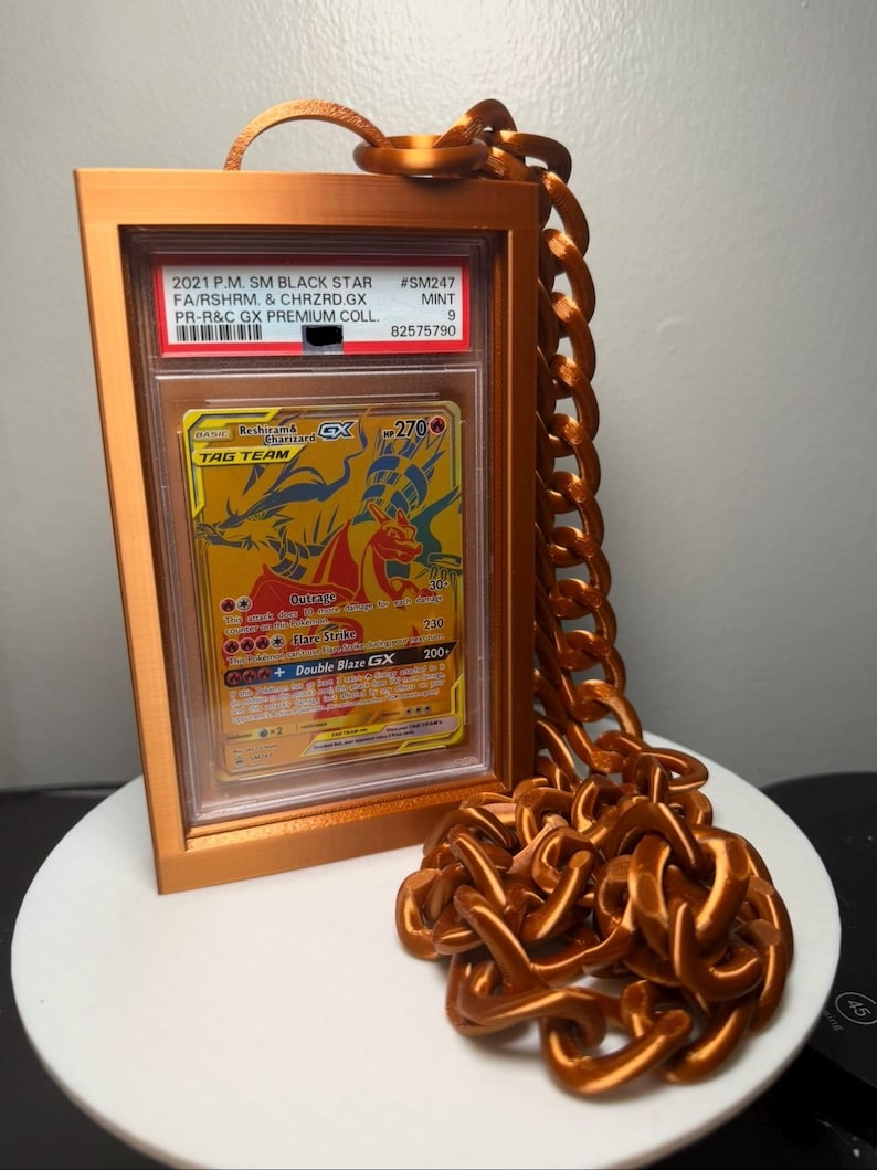 Peut inclure: Une carte &agrave; collectionner encadr&eacute;e avec un design dor&eacute; et orange, avec le texte "Reshiram & Charizard GX TAG TEAM". Le cadre et la cha&icirc;ne sont de couleur cuivre m&eacute;tallique. La carte est not&eacute;e et &eacute;tiquet&eacute;e avec le texte "2021 P.M. SM BLACK STAR".