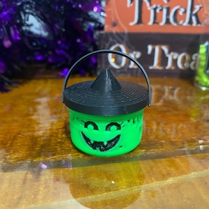 Puede incluir: Un caldero de Halloween verde con una tapa de sombrero de bruja negra y una cara sonriente. El caldero tiene un asa y está hecho de plástico.
