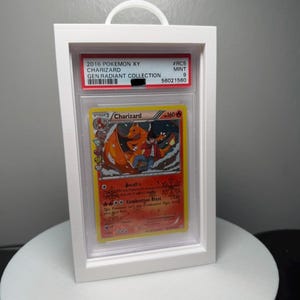 Peut inclure: Un pr&eacute;sentoir blanc encadr&eacute; contenant une carte Pok&eacute;mon Charizard de la collection Gen Radiant 2016. La carte pr&eacute;sente une illustration de Charizard orange flamboyant. Le bo&icirc;tier a une poign&eacute;e blanche en haut.