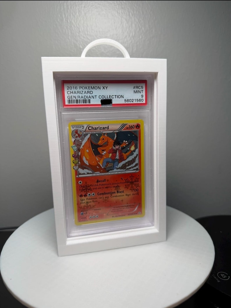 Peut inclure: Un pr&eacute;sentoir blanc encadr&eacute; contenant une carte Pok&eacute;mon Charizard de la collection Gen Radiant 2016. La carte pr&eacute;sente une illustration de Charizard orange flamboyant. Le bo&icirc;tier a une poign&eacute;e blanche en haut.