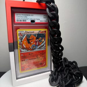 Peut inclure: Une carte Pok&eacute;mon encadr&eacute;e avec Charizard, avec un cadre rouge et blanc et une cha&icirc;ne noire. La carte provient de la collection 2016 Pok&eacute;mon XY Gen Radiant. La carte a une note de 9.