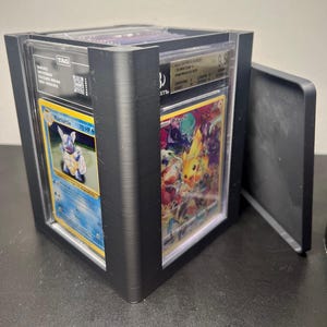 May include: Black display case holding two graded Pokémon trading cards. One card features a blue Wartortle, and the other showcases a colourful Pikachu. The case has a hinged door and a TAG label.