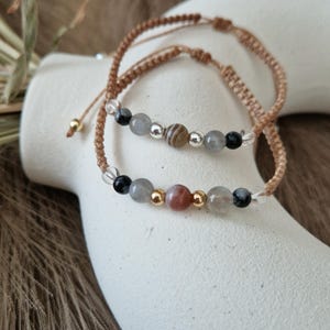 Könnte beinhalten: Zwei Armbänder mit braunem Band und Edelsteinperlen. Ein Armband hat eine hellrosa, graue und schwarze Perle. Das andere Armband hat eine braune, graue und schwarze Perle.