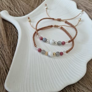Könnte beinhalten: Zwei geflochtene braune Armbänder mit Edelsteinperlen. Die Armbänder haben einen Mix aus hellgrauen, weißen und rosafarbenen Perlen. Die Armbänder werden auf einer weißen Keramikplatte präsentiert.