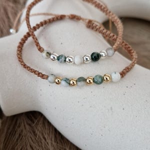 Könnte beinhalten: Zwei Makramee-Armbänder mit Natursteinperlen. Die Armbänder haben eine braune Kordel und eine Mischung aus weißen, grünen und silbernen oder goldfarbenen Perlen. Die Armbänder werden auf einer weißen Oberfläche präsentiert.