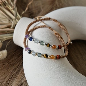 Könnte beinhalten: Zwei Armbänder mit braunem Band und bunten Edelsteinperlen. Das obere Armband hat blaue, grüne und silberne Perlen. Das untere Armband hat lila, grüne, orange und rote Perlen.