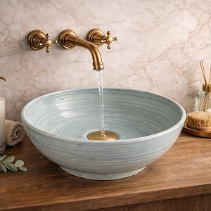 Puede incluir: Un lavabo de cerámica azul claro con un grifo de latón, un dispensador de jabón y accesorios. El lavabo está montado en un mueble de madera. El agua fluye del grifo al lavabo. El fondo es una pared de mármol.