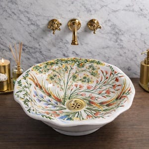 Puede incluir: Un lavabo de cerámica blanca con estampado floral y desagüe dorado, sobre una pared de mármol. Grifos dorados, un dispensador de jabón y una vela completan la decoración del baño. El lavabo presenta un colorido diseño botánico.