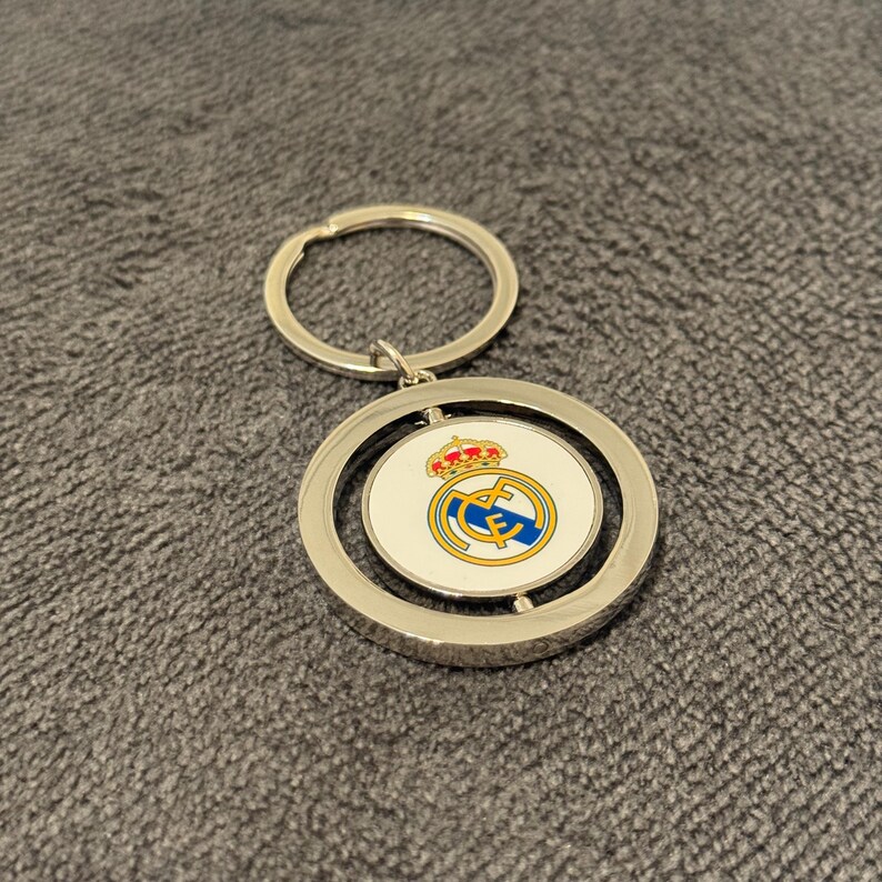 May include: Silver keyring with a circular pendant displaying the Real Madrid crest. The crest features a crown, a gold and blue design on a white background, and a silver key ring.