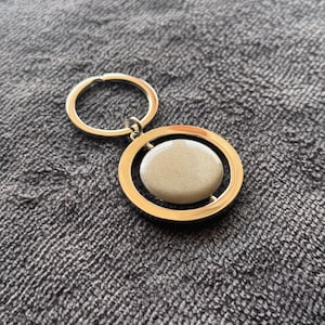 May include: A gold-coloured keyring with a circular design. The keyring features a gold ring and a pendant with a gold frame surrounding a light beige oval stone. The background is a grey textured fabric.