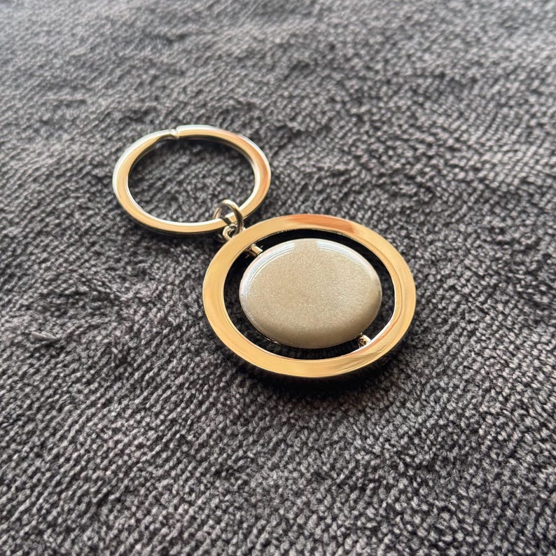 May include: A gold-coloured keyring with a circular design. The keyring features a gold ring and a pendant with a gold frame surrounding a light beige oval stone. The background is a grey textured fabric.