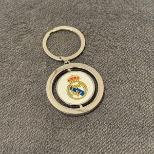 May include: A silver keyring with a circular pendant displaying the Real Madrid logo. The logo incorporates a crown, a blue and yellow design, and the team's initials. The pendant rotates and is attached to a silver key ring.